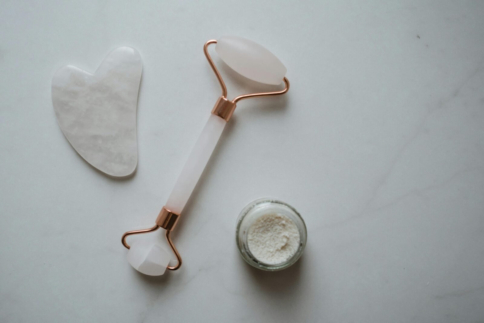 Flat lay of a gua sha, facial roller, and natural face scrub on a marble surface for skincare routines.